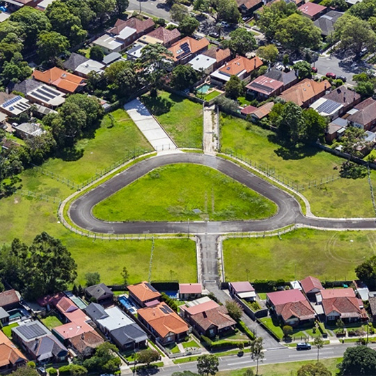 An aerial view of am empty grassy lot of land with a looping street running through it, surrounded on all sides by single-dwelling housing blocks.